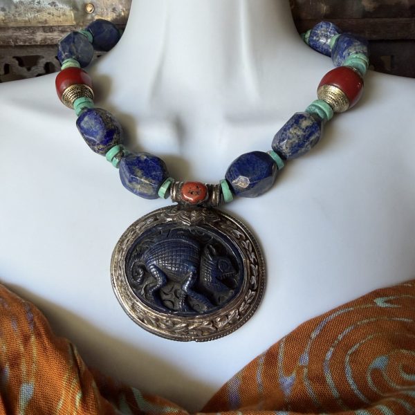 22. The Carved Blue Copal Pendant Necklace by artist Barbara Wright of Dallas, TX, features a beautiful pendant made from carved Mexican blue copal which depicts a chameleon . This pendant is complemented with beautiful turquoise, red copal (Tibet) , and lapis lazuli beads (Afghanistan,) measuring 18 inches in length. The design is inspired by several different world cultural traditions, showcasing intricate craftsmanship and a captivating bluish color palette. The pendant is made with Tibetan silver metal and sourced materials from Tibet