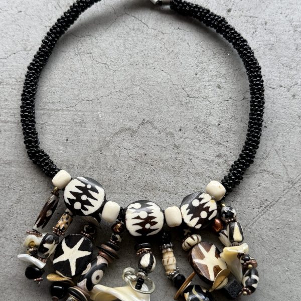 This stunning West African hand-dyed batik, bovine, lava rock, natural and mother-of-pearl beaded necklace designed by Jo Anne Brooks is a coversation-starter! The necklace features round-shaped beads made of black, brown, and white authentic materials ethically sourced from Kenya, showcasing a unique and ethnic regional style. The black and cream batik beads are crafted from bovine and the design is handmade, creating a visually striking piece that is sure to make a statement. Perfect for adding a touch of authentic African craftsmanship to any outfit, this necklace is a gorgeous and culturally-inspired accessory for any jewelry lover.